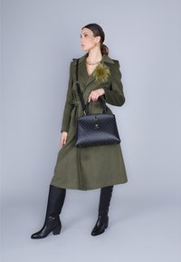 Trench coat verde oliva con cintura, borsa nera con pois bianchi e stivali neri al ginocchio con dettagli a bottoni.
