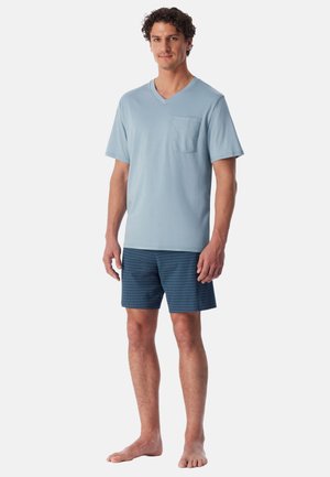 Man standing barefoot wearing a light blue V-neck short sleeve shirt with pocket and dark blue patterned shorts against a white background.