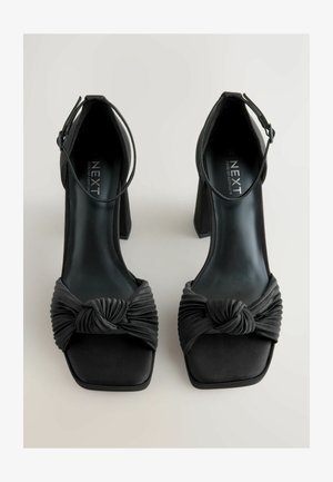 Črne visokokotne sandale s kvadratnim prstom, satenastim materialom, z vozlasto tkanino na sprednji strani in z naramnico okoli gležnja za podporo.