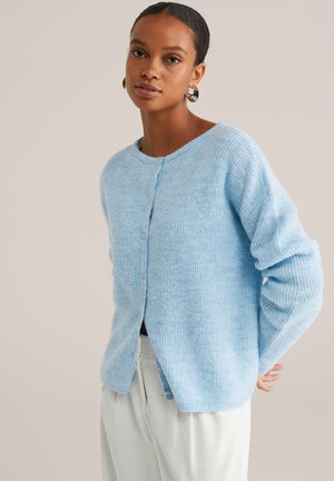 Femme aux cheveux bruns portant un cardigan bleu clair boutonné et un pantalon blanc, avec des boucles d'oreilles créoles, se tenant devant un fond clair simple.