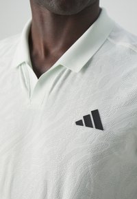 Polo shirt vert clair avec un motif texturé. Présente un logo Adidas noir sur le côté gauche de la poitrine.