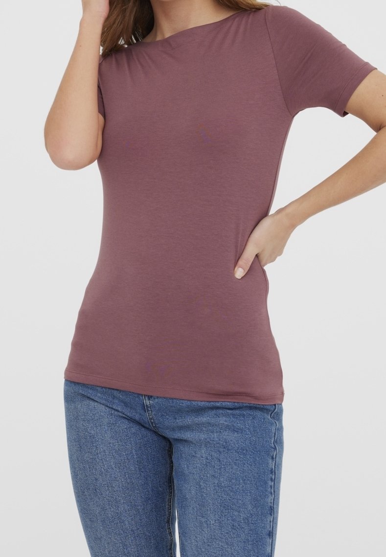 Femme portant un t-shirt mauve à manches courtes et un jean bleu, une main sur la hanche et l'autre touchant ses cheveux.