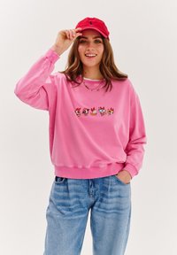 Różowy sweter z prążkowanymi mankietami i brzegiem, z kolorową haftowaną postacią z kreskówki. Połączony z jasnoniebieskimi, luźnymi dżinsami.