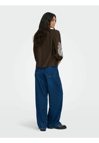 Brun, struktureret, cropped top med puffede ærmer, der har hvid blomstermønster broderet på venstre ærme; parret med løse blå denim bukser.
