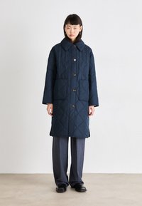 Manteau matelassé bleu marine avec un grand col, fermeture à boutons, deux poches avant et fentes latérales pour faciliter les mouvements. Design de surface texturé.