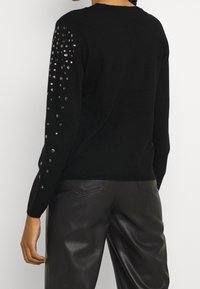 Pull noir à manches longues orné de sequins argentés sur la manche gauche ; assorti à un pantalon en cuir noir.