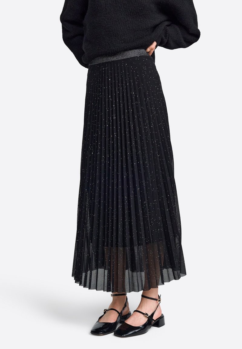 Jupe plissée noire avec une couche transparente et des paillettes métalliques ; dotée d'une large ceinture et associée à des chaussures en cuir verni.