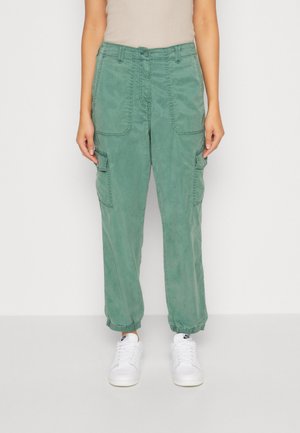 Groene cargo-broek met grote zijzakken, een losse pasvorm en elastische boorden, gemaakt van een zachte, gestructureerde stof. In combinatie met witte sneakers.