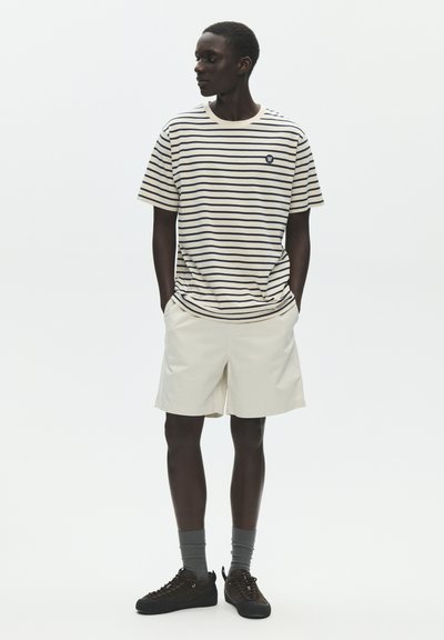 Homme portant un t-shirt à rayures crème et marine, un short beige clair, des chaussettes grises et des baskets noires, debout les mains dans les poches regardant vers la gauche.