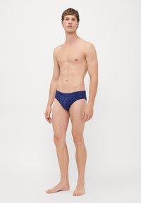 Junger Mann steht barfuß und trägt blaue Schwimmtrunks mit einem schwarzen Allover-Dsquared2-Logo-Muster auf weißem Hintergrund.