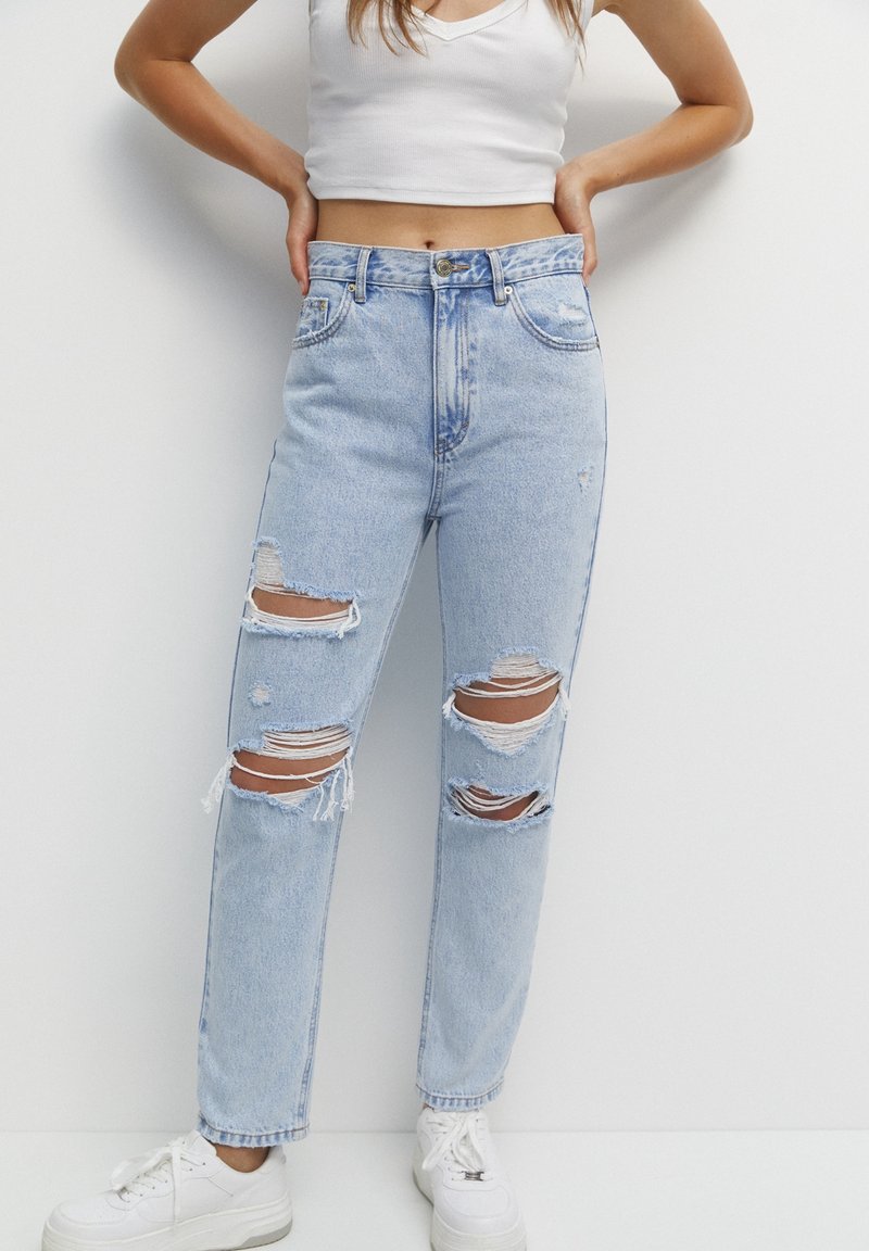 Femme portant un jean déchiré bleu clair, un crop top blanc et des baskets blanches, debout les mains sur les hanches sur un fond uni.