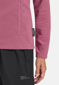 Giacca in pile rosa con una texture morbida, dotata di un'etichetta Polartec sul lato e una tasca visibile. Pantaloni neri con logo.