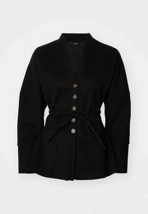 Cardigan noir avec décolleté en V, fermeture à boutons et ceinture à nouer. Il présente une texture douce et des manches larges, créant une silhouette décontractée.