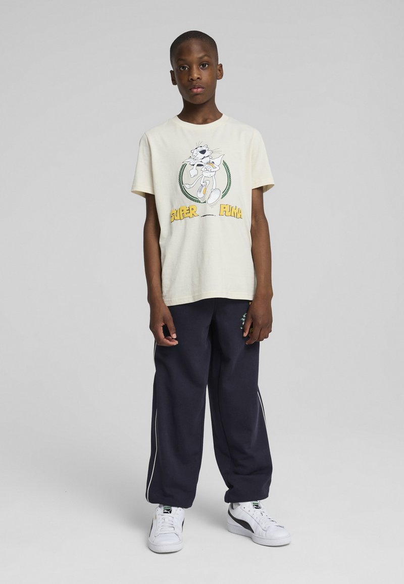 T-shirt in cotone crema con grafica di cartone animato, abbinata a pantaloni felpati larghi color navy con strisce laterali bianche e sneakers bianche.