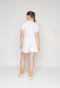 Hvit skjorte med korte ermer og shorts, med en avslappet passform. Orange sokker med chevronmønster. Modell vendt bort, på en nøytral bakgrunn.