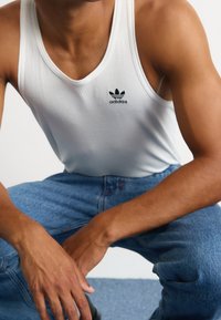 Top sin mangas blanco de canalé con un logo negro de Adidas, combinado con unos jeans de mezclilla azules. La tela tiene una apariencia texturizada, mostrando un diseño casual.