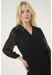 Schwarzes Kleid mit transparenten langen Ärmeln, V-Ausschnitt und plissiertem Rock. Der Stoff fällt weich und schafft eine fließende Silhouette.