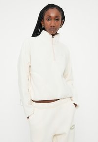 Pullover in pile di colore avorio con colletto a zip, vestibilità rilassata e polsini a costine. Abbinato a pantaloni della tuta rilassati in coordinato con testo verde sobrio.