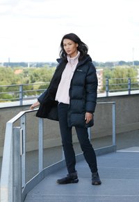 Moteris juodame pūkuotame džemperyje ir juodose kelnių stovinti ant metalinės tako, laikydama turėklą, o fone matosi medžiai ir pastatai.