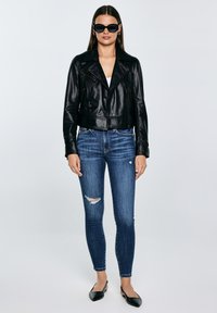 Moteris juodos odinės striukės, suplėšytų siaurų mėlynų džinsų, juodų smailių batelių ir didelių juodų akinių nuo saulės, stovinti priešais vientisą foną.