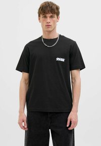 Schwarzes Baumwoll-T-Shirt mit kurzen Ärmeln, das ein weißes Logo auf der linken Brust zeigt. Kombiniert mit einer dunklen Jeans und einer silbernen Kettenhalskette.