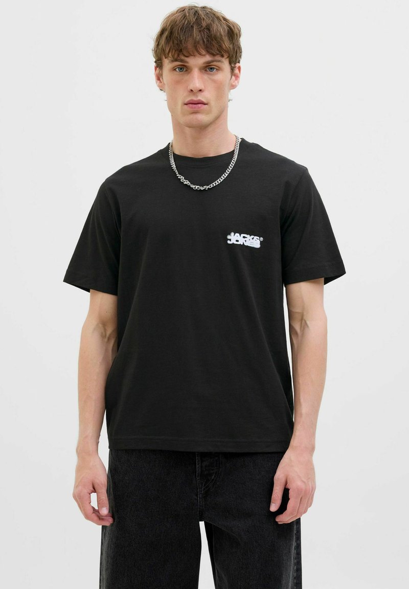 Schwarzes Baumwoll-T-Shirt mit kurzen Ärmeln, das ein weißes Logo auf der linken Brust zeigt. Kombiniert mit einer dunklen Jeans und einer silbernen Kettenhalskette.