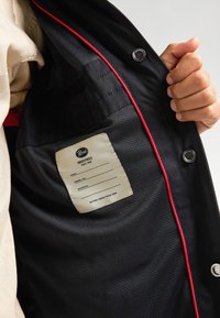 Interiorul unei jachete negre prezentând o căptușeală din plasă, cusături roșii și un etichetă cu numele mărcii și un spațiu pentru detalii personale. Un buton cu cap tăiat metalic este vizibil.