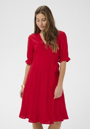 Žena s dlhými vlnitými vlasmi, ktorá má na sebe červené zavinovacie šaty s rukávmi do lakťov a volánovým lemom, pozerá sa dolu s jemným úsmevom.