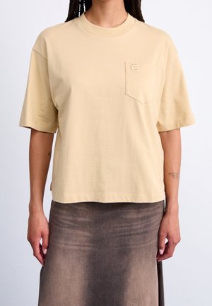 Personne portant un t-shirt beige à manches courtes avec une poche sur la poitrine et une jupe marron foncé délavée, debout devant un fond blanc uni.