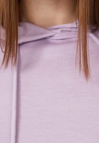Joven con cabello recto castaño claro, vistiendo una sudadera ligera color púrpura con cordones, visible desde los hombros hasta el cuello.