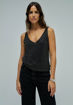 Femme aux longs cheveux bruns portant un haut noir sans manches à pois blancs, un pantalon noir et un bracelet argenté, debout devant un fond gris uni.