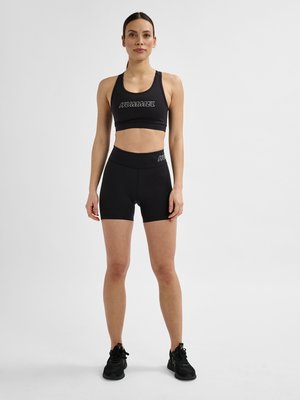 Vrouw staand, draagt een zwarte sportbeha en bijpassende zwarte shorts met hoge taille met merkslogo's, zwarte sportschoenen, neutrale achtergrond.