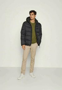 Μαύρο μπουφάν τύπου puffer με κουκούλα, με επένδυση σε καπιτονέ σχέδιο. Φορεμένο πάνω από πράσινο πουκάμισο, συνδυασμένο με μπεζ παντελόνι και λευκά αθλητικά παπούτσια.