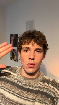 Ung mand med krøllet hår og en ørering i hoop-stil iført en stribet sweater, der løfter hånden tæt på ansigtet i et rum med vægplakater.