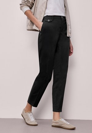 Person iført korte sorte bukser, beige bomberjakke, hvid skjorte og beige sneakers, stående mod en lyserød væg.