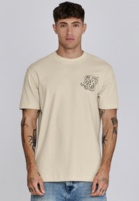 Beige Baumwoll-T-Shirt mit einem kleinen schwarzen Grafik auf der linken Brust. Kurze Ärmel und runder Halsausschnitt. Weiche Textur und lockere Passform.