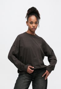 Σκούρο καφέ φαρδιά μπλούζα με μακριά μανίκια, λεπτομέρειες με ριμπ μανσέτες και άνετη εφαρμογή, σε συνδυασμό με σκούρο τζιν. Μινιμαλιστικός σχεδιασμός.