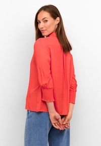 Camicia a maniche lunghe color corallo con polsini arricciati e vestibilità rilassata, abbinata a jeans in denim azzurro chiaro. Il tessuto è liscio e leggero.