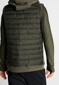 Uomo che indossa un gilet imbottito a quadri verde scuro sopra una maglietta a maniche lunghe testurizzata con cappuccio, visto da dietro su uno sfondo semplice.