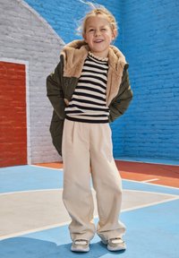 Jong meisje met een glimlach, met handen achter haar rug, gekleed in een gestreepte trui, groene jas met bontvoering, beige broeken en sneakers op een kleurrijk sportveld.