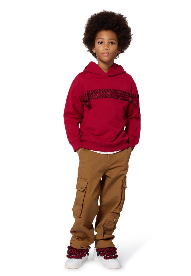 Felpa rossa con una striscia nera decorata sull'area del petto, pantaloni cargo beige e scarpe da ginnastica bianche con dettagli rossi e polsini testurizzati.