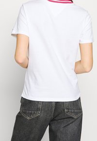 Camiseta blanca de algodón con un collar de rayas rojas y negras, de mangas cortas y corte clásico, combinada con unos vaqueros oscuros de cintura alta.