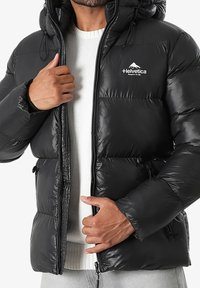 Homme portant une veste en duvet noire avec fermeture éclair et un pull en tricot blanc en dessous, ajustant le devant de la veste près du logo sur la poitrine.