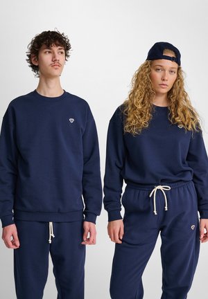 To unge voksne iført matchende marineblå hættetrøjer og joggingbukser, står side om side mod en enkel lys baggrund.