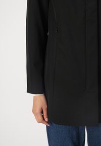 Manteau noir avec un col haut et une coupe droite. Il dispose d'une poche zippée, de manches longues et est en tissu lisse. Un jean bleu est visible.