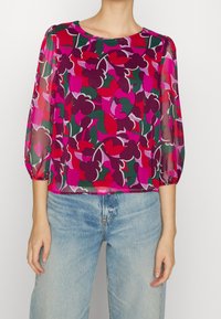 Blouse longue à manches longues en tissu transparent avec un motif floral rose, rouge et vert. Encolure ronde et manches avec poignets élastiques, portée avec un jean bleu clair.