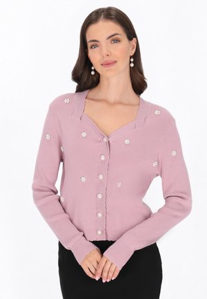 Vrouw draagt een lichtpaarse cardigan met parelknopen en kleine bloemversieringen, gecombineerd met een zwarte rok.