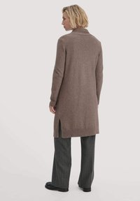 Cardigan lungo marrone in morbido knit, con colletto, spacchi laterali e polsini a coste. Abbinato a pantaloni grigi a righe.