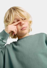 Mintgrøn sweatshirt med ribbet krave og cuffs, lavet af blødt stof. Ung person rører ved deres ansigt, med uordnet blond hår.