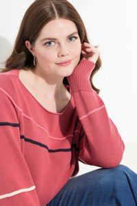 Różowy sweter robiony na drutach z prążkowanymi mankietami, z białymi i granatowymi poziomymi paskami, zestawiony z niebieskimi dżinsami. Modelka stoi w pozie z ręką blisko twarzy.